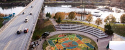 Aerial view of a riverside amphitheater with colorful painted ground designs, near a bridge with light traffic and trees with autumn foliage.