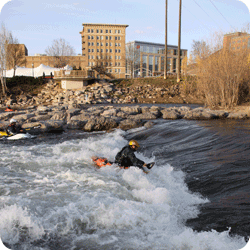 A person in a helmet and life jacket kayaks through whitewater rapids, with buildings and trees visible in the background.