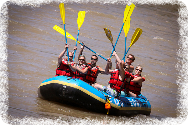 Group Programs 8 Six people wearing red life jackets hold yellow paddles up while sitting in a blue inflatable raft on brown river water.