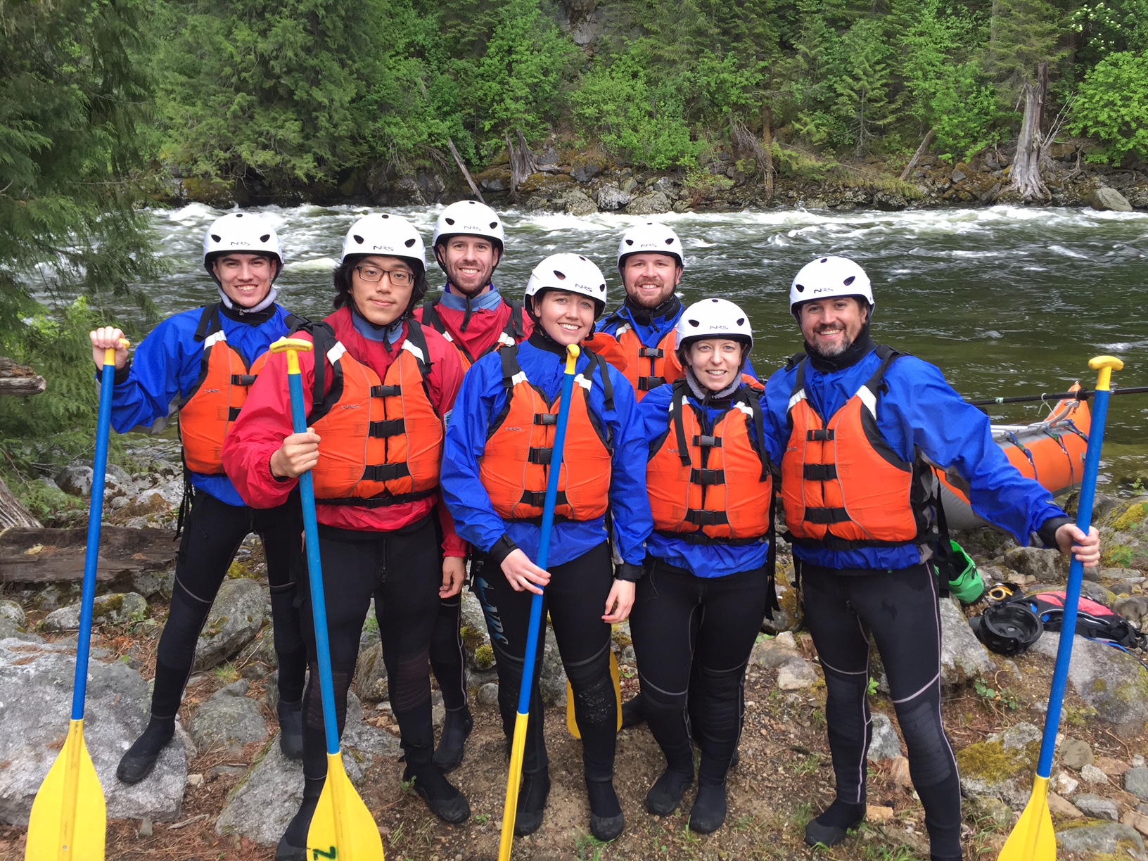 Eight people wearing helmets and life jackets stand together with paddles by a river, surrounded by trees, preparing for rafting.