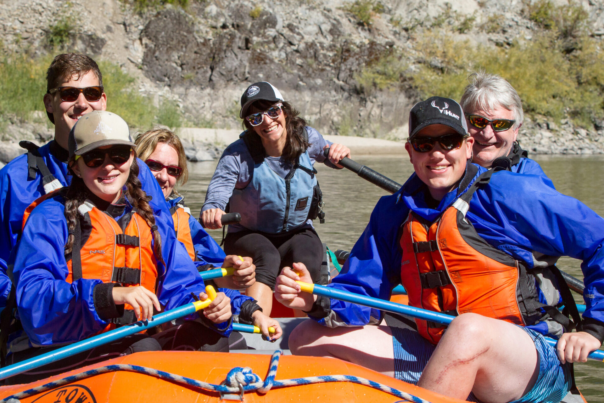 Six people wearing life jackets and sunglasses are smiling while rafting together on a river, enjoying an exciting rafting guide experience with rocky terrain in the background.