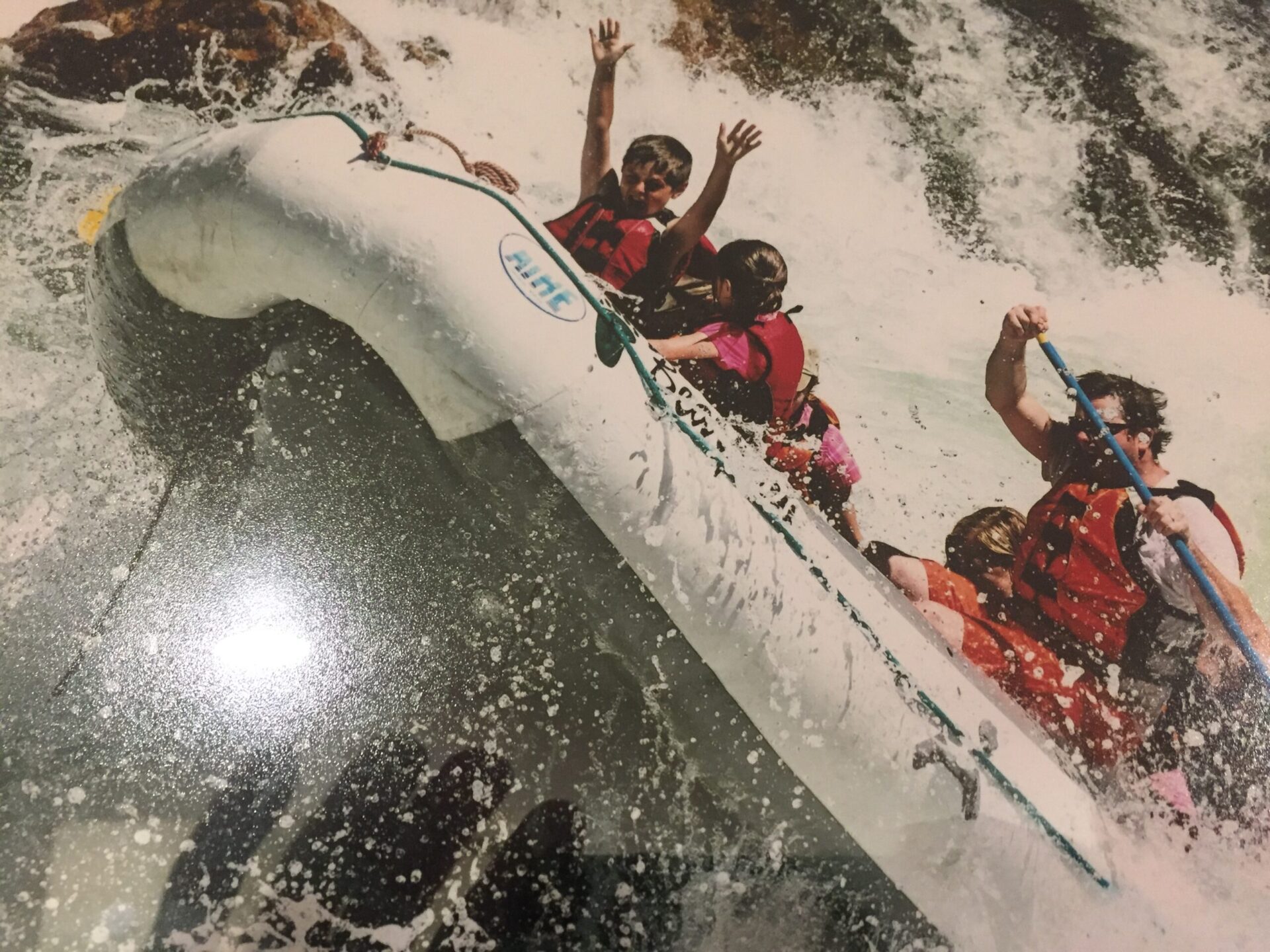 A group of people, including children in life jackets, enjoy rafting with kids as they ride a white inflatable raft through rough, splashing rapids.