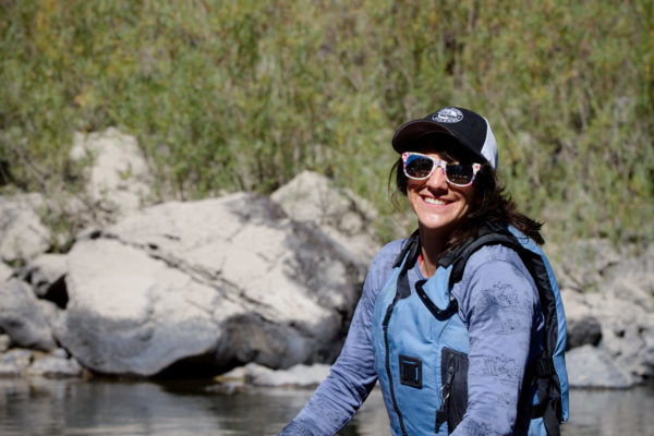A woman’s guide to river life: A woman wearing a life jacket, baseball cap, and sunglasses stands near a river, with rocks and bushes in the background.