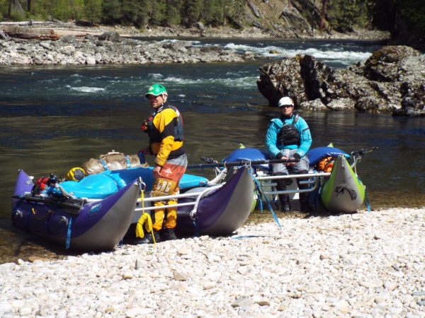 Two people wearing helmets and life jackets stand next to two inflatable rafts on a rocky riverbank with a forested river in the background.