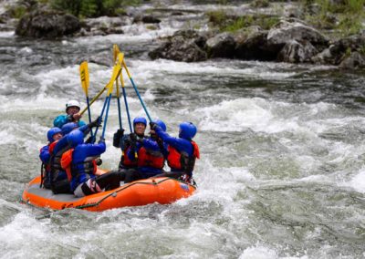 A group of rafters in blue helmets and life jackets raise their paddles together while navigating the rapids of the Lochsa River.