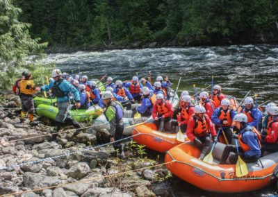 A group of rafters in helmets and life jackets prepares to launch on a guided rafting trip along the riverbank.