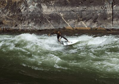 A person in a wetsuit surfs on a river wave near a rocky shoreline.