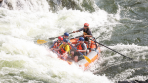A group of people wearing helmets and life jackets paddle an inflatable raft through whitewater rapids.