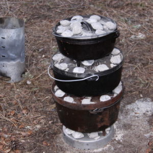 Three cast iron Dutch ovens are stacked outdoors, each topped with charcoal briquettes for cooking. A metal chimney starter sits on the ground nearby.