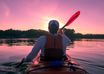 Kayaking 7 A tourist is wearing safety gear and paddling in a kayak on the blackfoot river. They are on a guided trip with Zoo Town Surfers. There is a stunning purple and pink sunset.