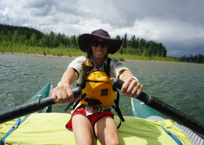 Private River Training with a boating learning to run the oars. She has a green cat raft, purple sun hat, yellow pfd safety vest and red shorts. She is holding the oars and behind her is green river water, a forest of trees on the shoreline and moody grey storm clouds.