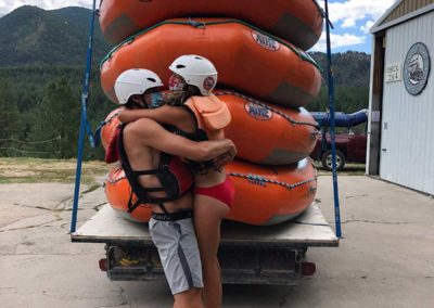 Ecstatic boaters embrace in a hug while wearing swim trunks, bikini, helmets and PFD at Zoo Town Surfers headquarters on the Alberton Gorge. There is a stack of 5 orange rafts behind them.