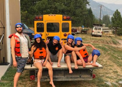 5 Zo Tow Surfers guides are seated on a raft trailer behind our shuttle bus outside of headquarters. They are smiling and wearing safety gear including blue helmets and orange PFDs.