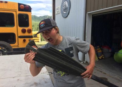 A Zoo Town Surfers Guide jokingly takes a bit out of a paddle that has a missing chunk. She is wearing a black hat with the Zoo Town Surfers logo on it and standing out front of our headquarters on the Alberton Gorge.