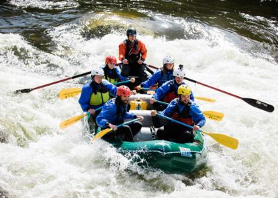 A Zoo Town Surfers raft is gliding over white water on the Lochsa River. Guests are holding yellow paddles and smiling while wearing drysuits and safety gear.