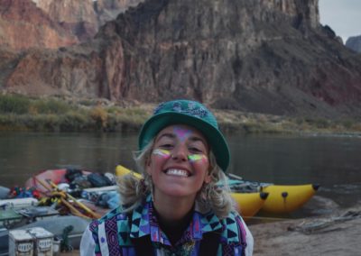 Overnight Trips 7 Abby a raft guide is smiling on a grand canyon adventure. Her face is painted and she has a colorful jacket and hat on.