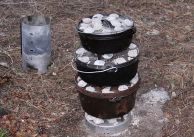 A camp stove with 3 dutch ovens and charcoal bricks is cooking a camp meal at a river campsite.
