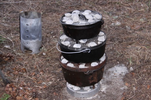A camp stove with 3 dutch ovens and charcoal bricks is cooking a camp meal at a river campsite.