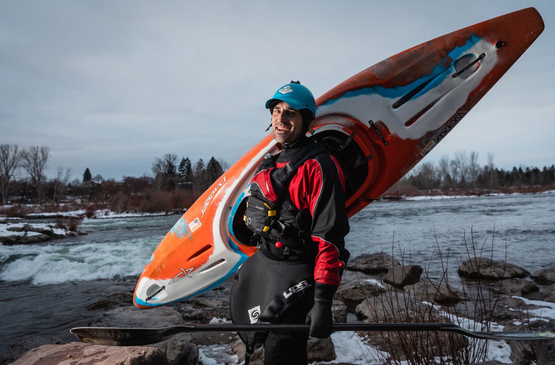A person wearing kayaking gear stands on rocks by a river, carrying an orange kayak on their shoulder and holding a paddle.