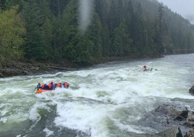 Adventurous whitewater rafting on the Lochsa River with Zoo Town Surfers. Rafters in bright safety gear navigate through intense rapids, surrounded by a dense forest and misty atmosphere. Zoo Town Surfers safety boat (a white cat raft) can be seen in the background. There is a hydraulic feature to the right where the rapid is churning water rapidly. To the right you can see the Selway-Bitterroot forest.