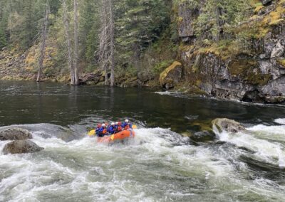 Whitewater rafting on the Lochsa River with Zoo Town Surfers. A group of rafters in bright safety gear paddles through a scenic section of the river, surrounded by rocky cliffs and lush forest. The image captures the beauty and adventure of rafting in this natural setting. The white water swirls different colors of green and you can see the mossy green and gold shores of the selway-bitterroot forest in idaho along the shoreline.