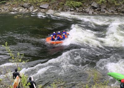 Whitewater rafting on the Lochsa River in Idaho with Zoo Town Surfers. A group of rafters in bright safety gear navigates through the river's rapids, surrounded by rocky terrain and lush greenery. Kayakers can be seen on the riverbank, adding to the adventurous scene.