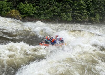 Adventurous whitewater rafting on the Lochsa River with Zoo Town Surfers. Rafters in vibrant safety gear tackle powerful, churning rapids, with water splashing dramatically around them. The lush forest backdrop emphasizes the thrill and natural beauty of the rafting experience.