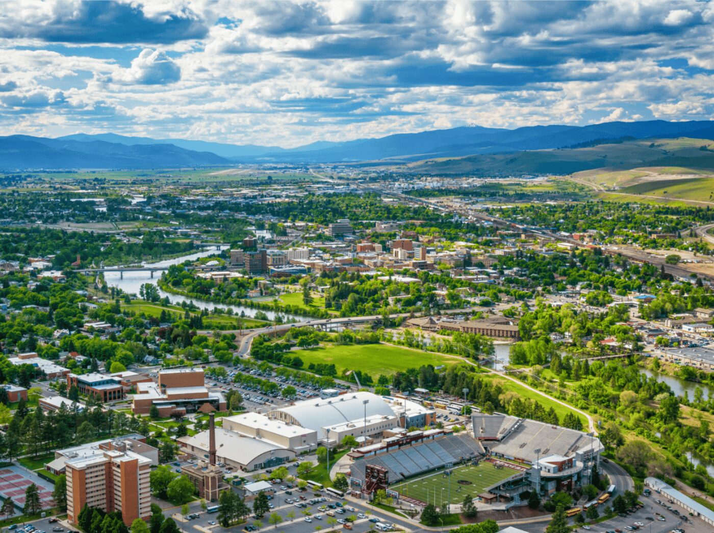 Things To Do In Missoula, MT Overlooking Missoula, MT on a sunny day there is blue sky with clouds in the background, a river runs through it, the University of Montana is pictured in the bottom left and there is a bustling downtown section with housing around it.