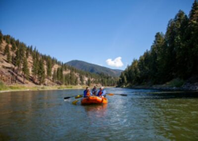 Learn To Series 7 A group of people paddles an orange inflatable raft down a river surrounded by forested hills and mountains under a clear blue sky.